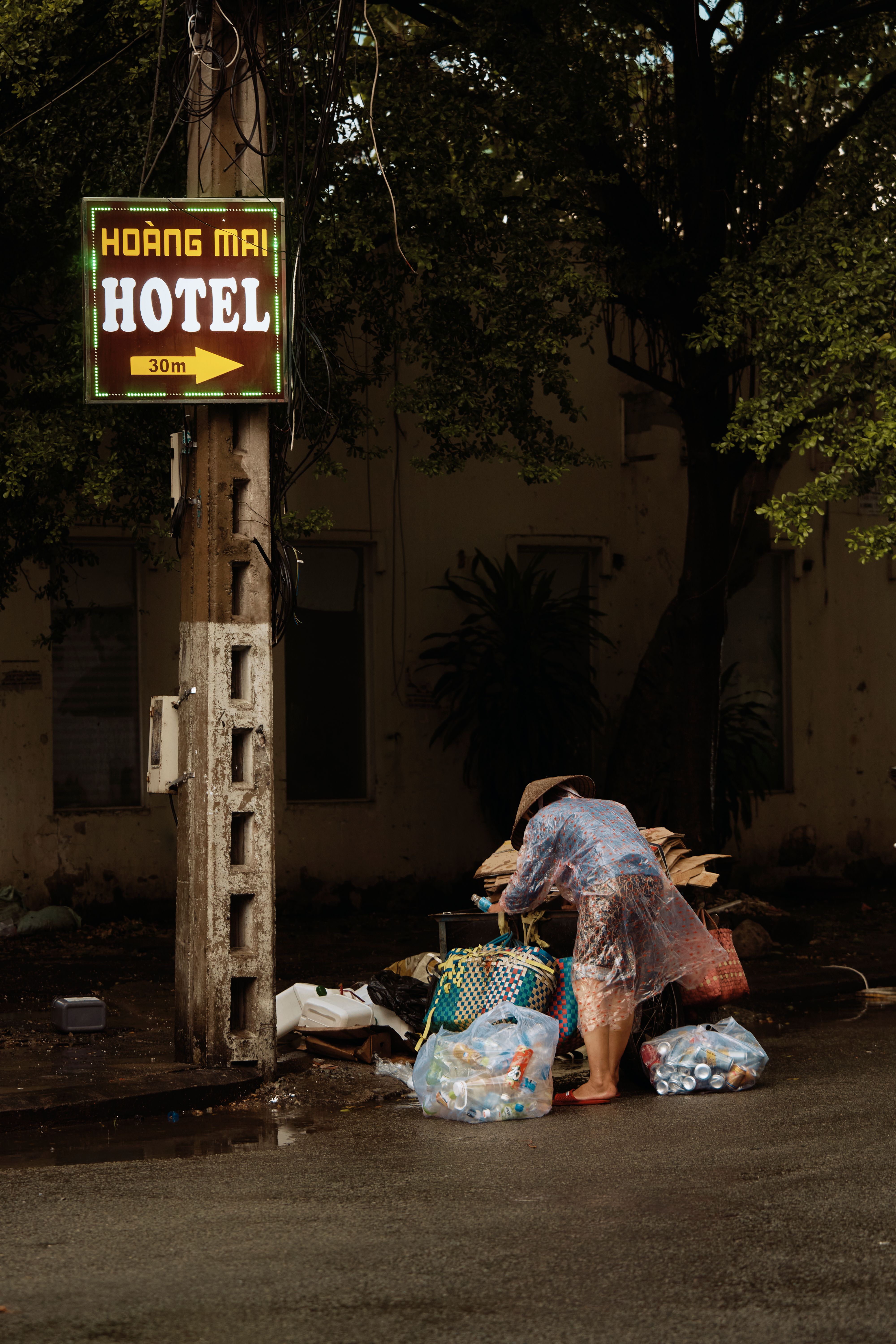 Person collecting recyclables in a rainy urban setting near Hoàng Mai Hotel sign.
