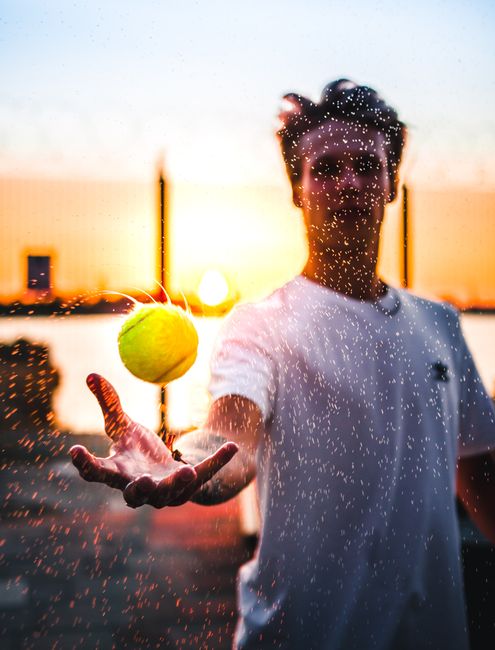 Vibrant photo of a man tossing a tennis ball, surrounded by water droplets at sunset in Tallinn.