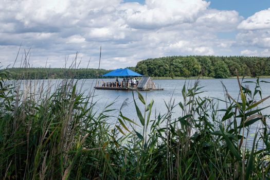 Scenic summer view of a lake with a floating raft under a blue umbrella surrounded by reeds.