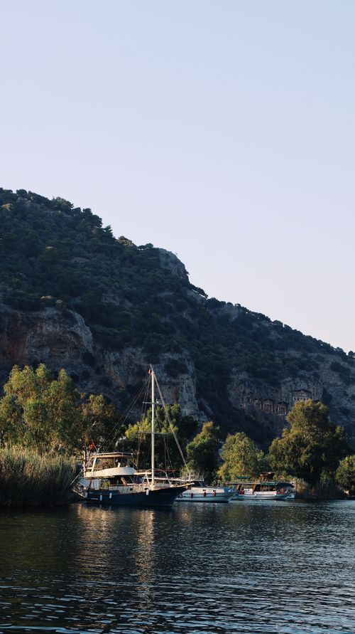 Free A serene view of a boat docked by a rocky shoreline surrounded by lush greenery at dusk. Stock Photo