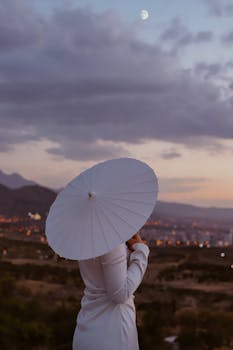 Woman in white dress with umbrella overlooking city at sunset.
