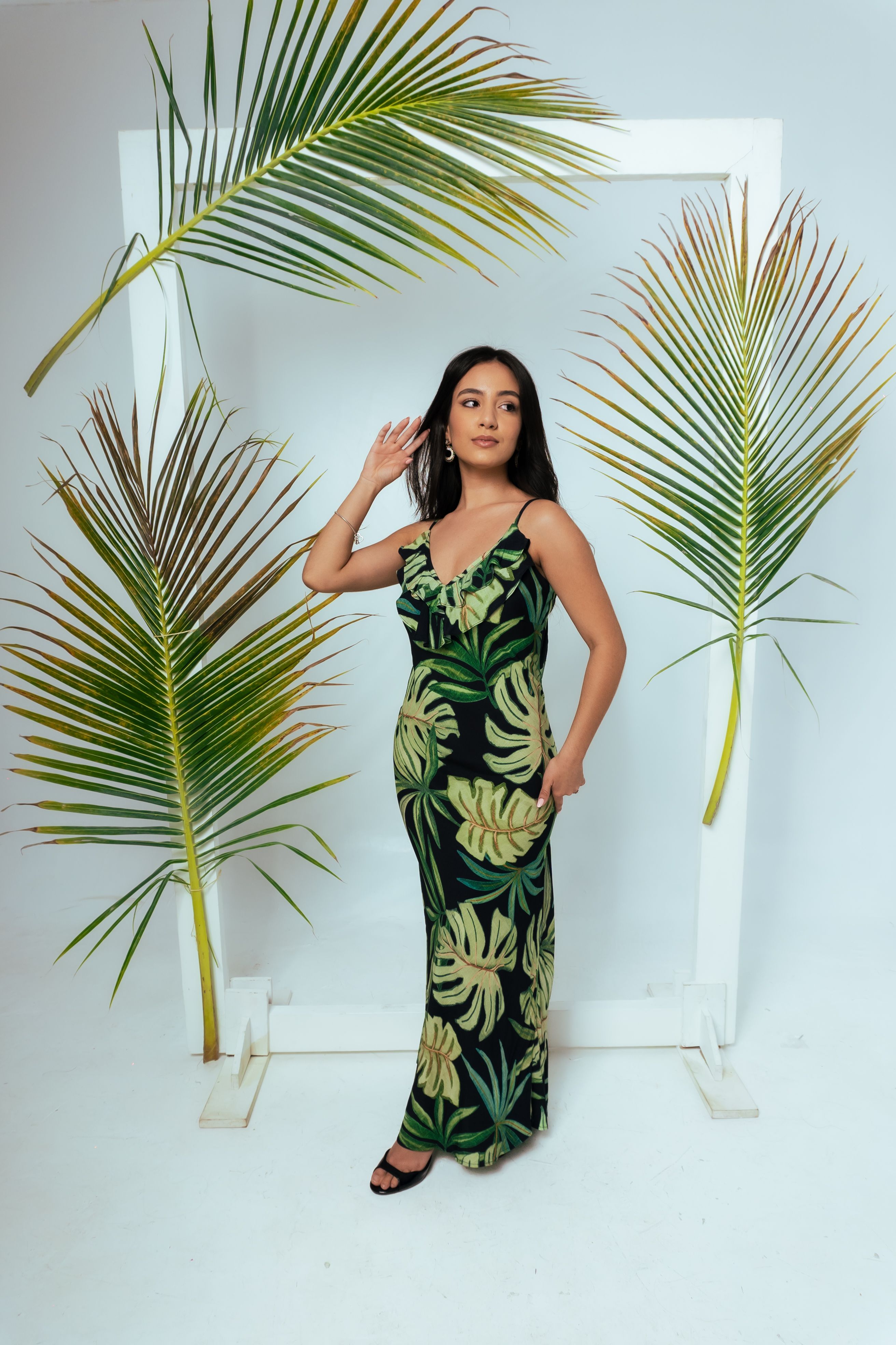 Woman in a tropical-themed dress poses elegantly among lush palm leaves indoors.