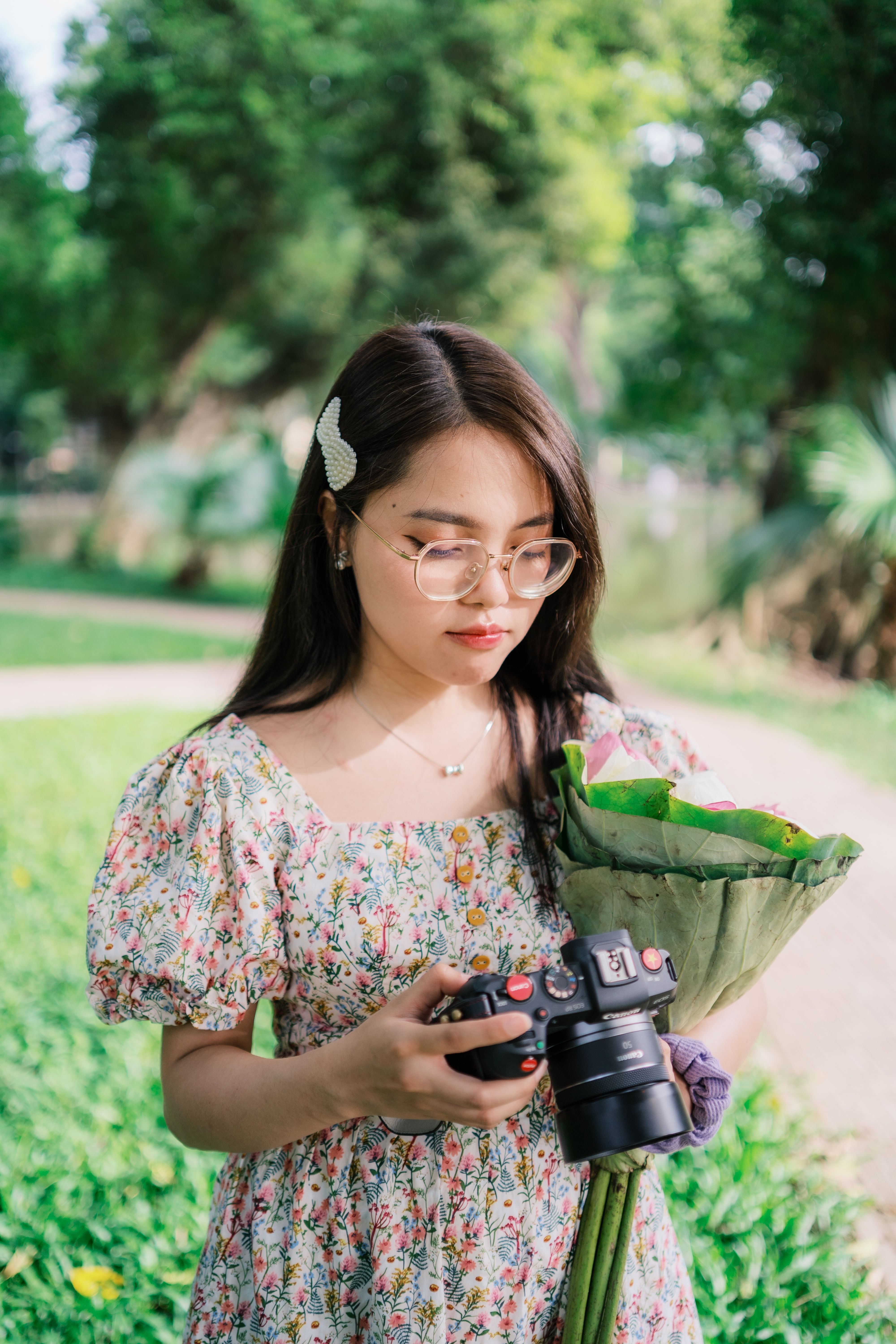 A young woman holding a camera and bouquet in a vibrant park, enjoying a sunny day.