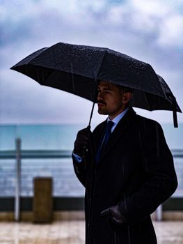A stylish man in a suit holds an umbrella by a rainy beach, exuding elegance.