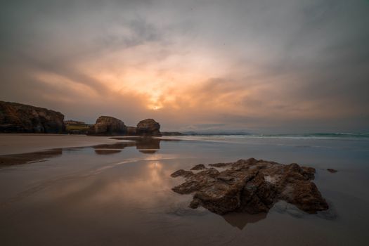 Tranquil beach scene with a serene sunset reflected on wet sand and rocks.
