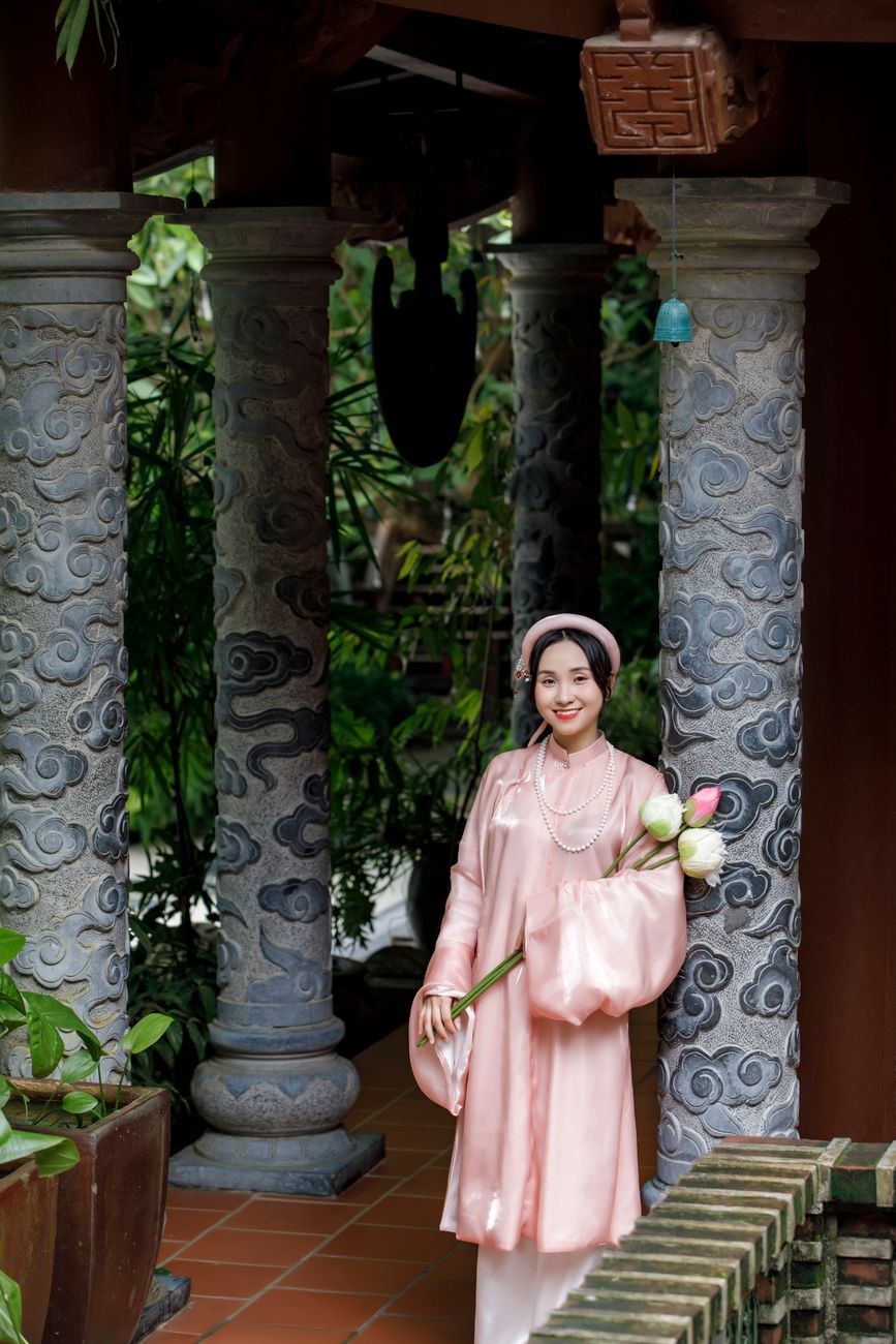 A woman wearing a traditional Vietnamese dress stands in an ornate garden setting, holding flowers.