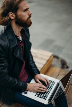 一个留着大胡子的男人专注地坐在长凳上，用笔记本电脑在户外远程工作