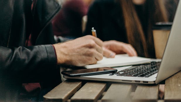 A freelancer writing notes in a notepad beside a laptop and smartphone on a desk.