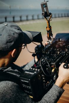 A man with a cap and eyeglasses operates a professional video camera during an outdoor shoot.