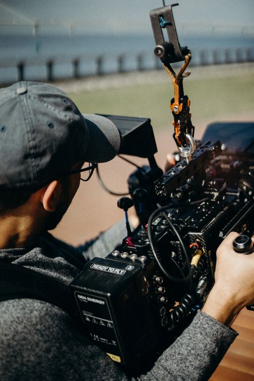 Free A man with a cap and eyeglasses operates a professional video camera during an outdoor shoot. Stock Photo