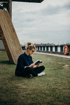 女人坐在水边的草地上看书，营造出一片宁静的氛围