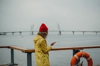 一名穿着黄色雨衣的妇女站在海滨栏杆旁，用智能手机拍照，背景是一座桥