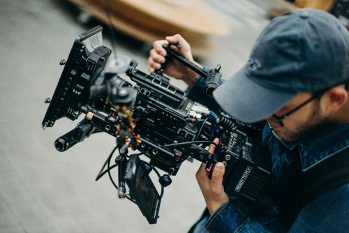 Free A man with a cap operates a professional video camera outdoors during daytime, capturing cinematic footage. Stock Photo