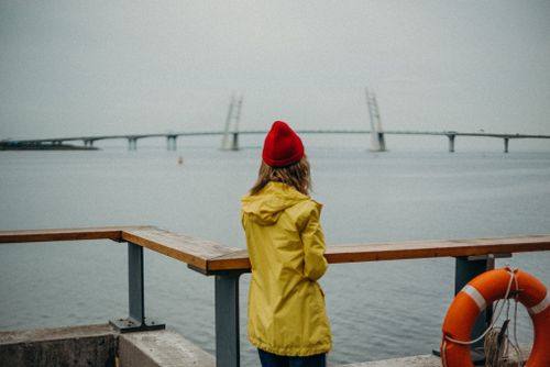 Free A woman in a yellow raincoat stands by the water, looking at a distant bridge. Stock Photo