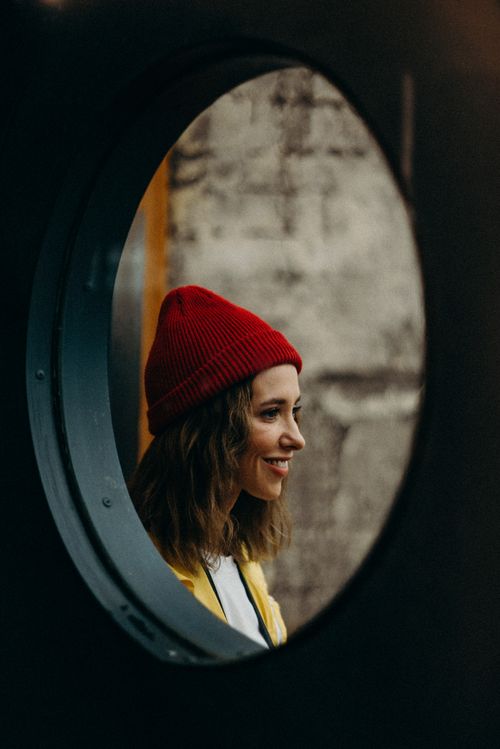 Free A woman in a red beanie smiles while looking through a circular opening, set against a rustic background. Stock Photo