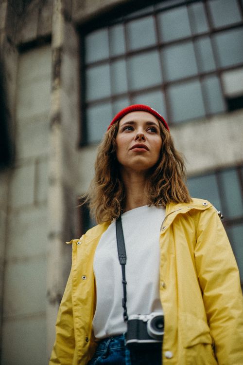 Free A young woman in a yellow jacket and red beanie poses outdoors with a camera, embodying urban style. Stock Photo
