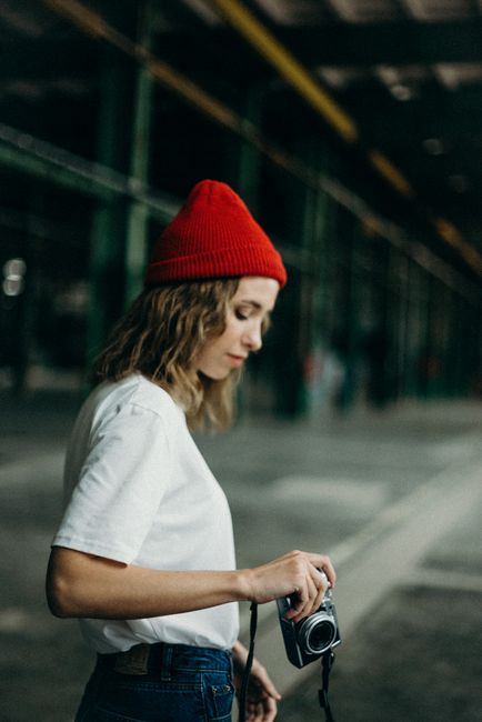 A young woman in a red beanie holding a retro camera, standing indoors with blurred background.
