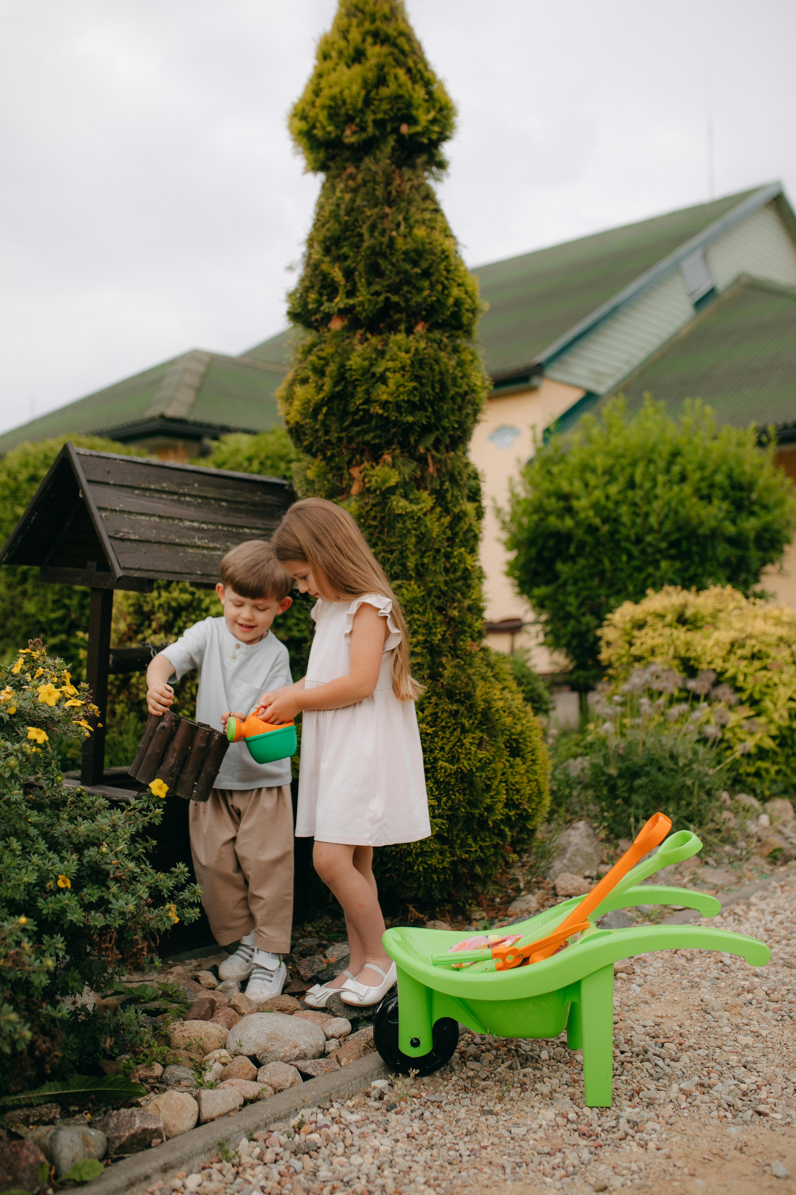 Two kids having fun outdoors, enjoying a summer day in a garden with a green wheelbarrow and toy tools.