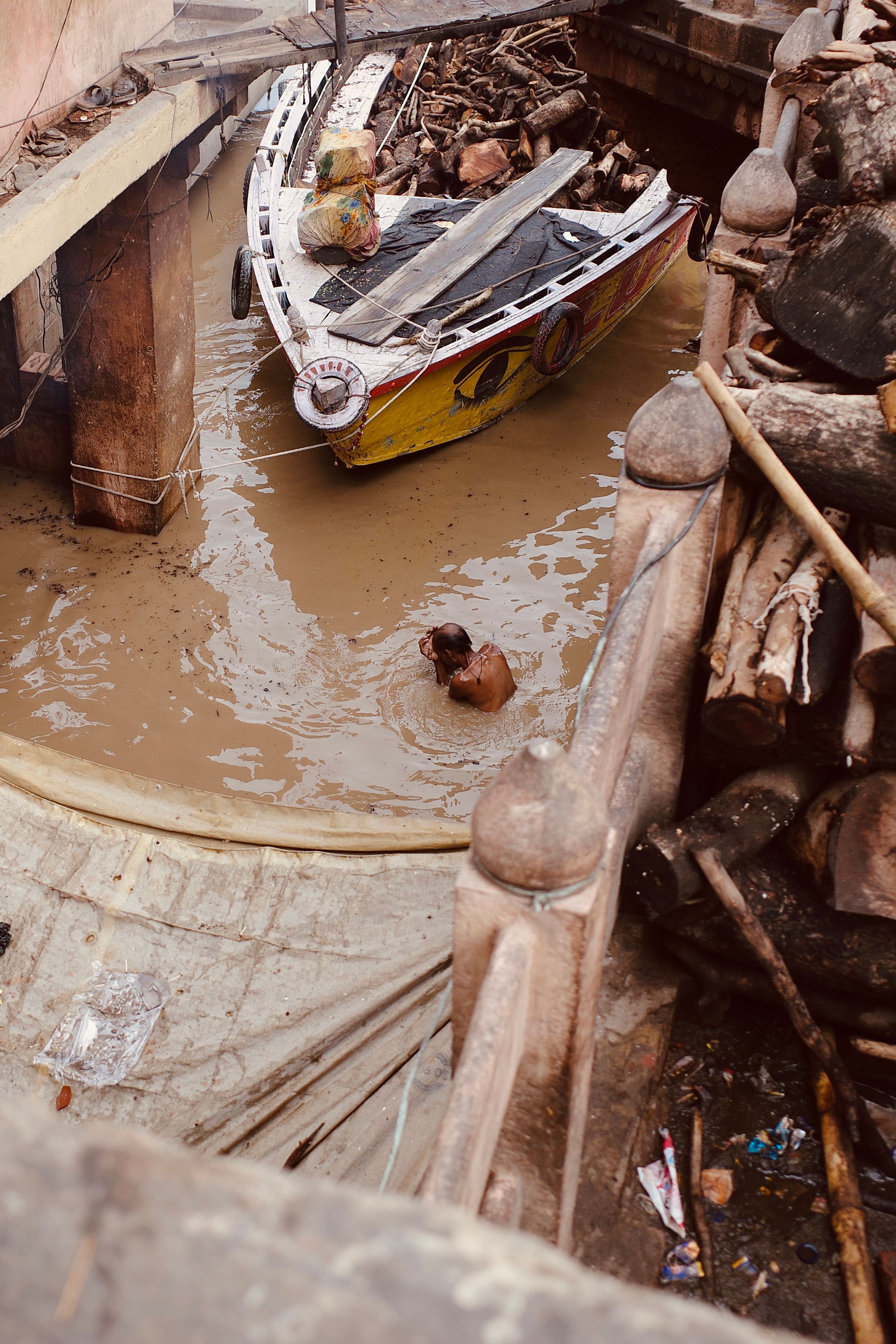 An overhead view of a traditional boat in a muddy river with a person bathing, amid scattered debris.