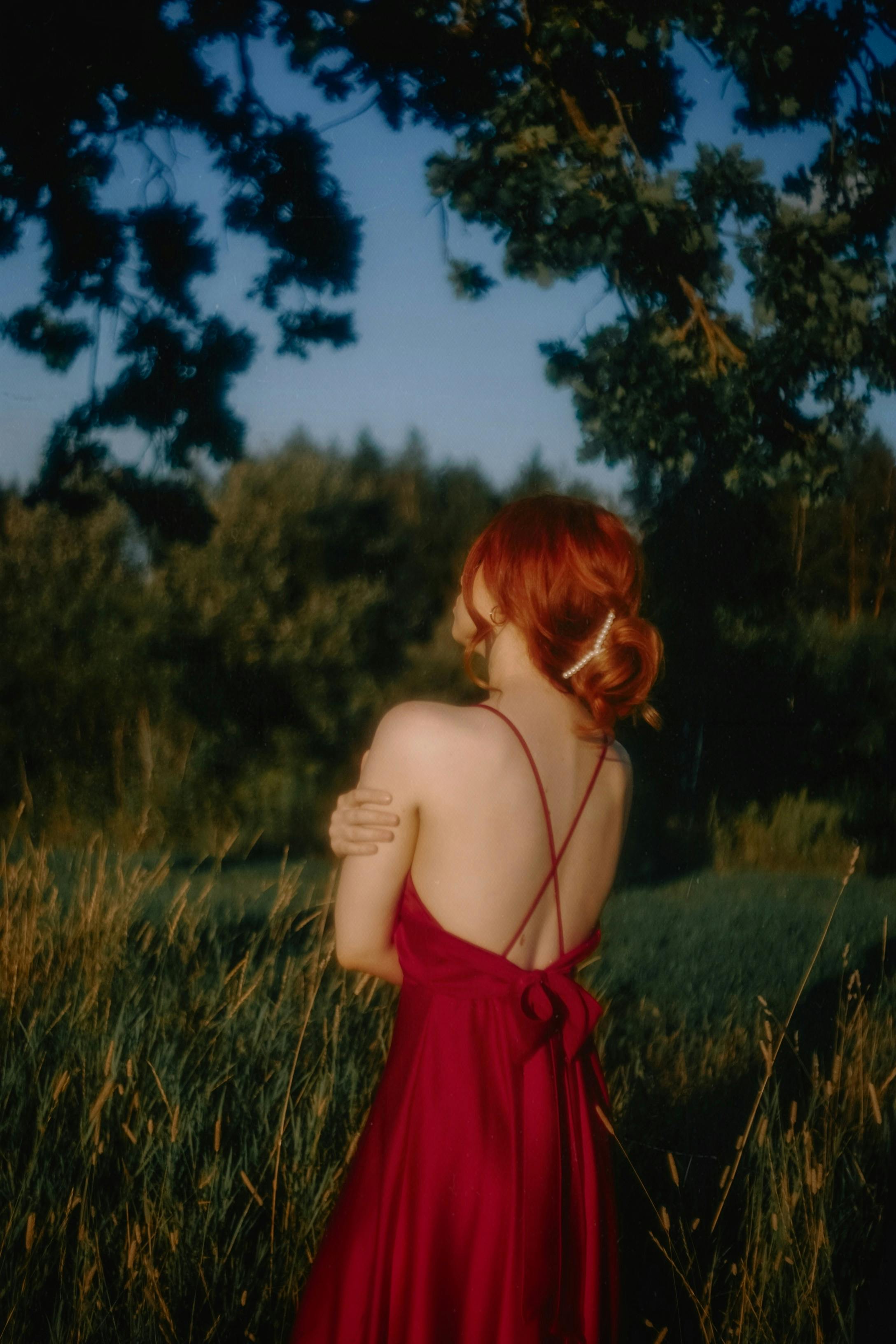 Woman in backless red dress standing in a serene, grassy meadow at dusk.