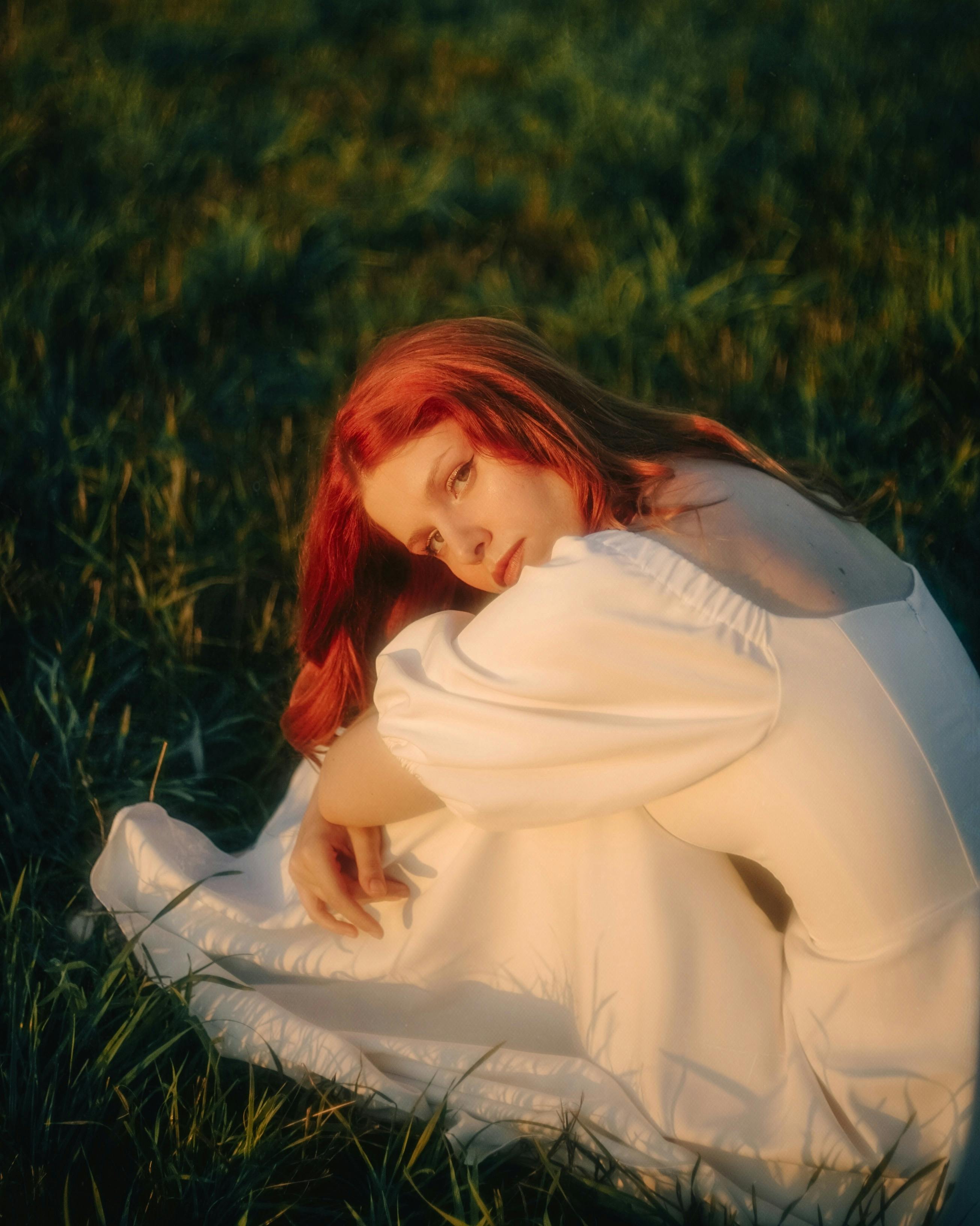 Portrait of a redhead woman in a white dress sitting gracefully on grass.