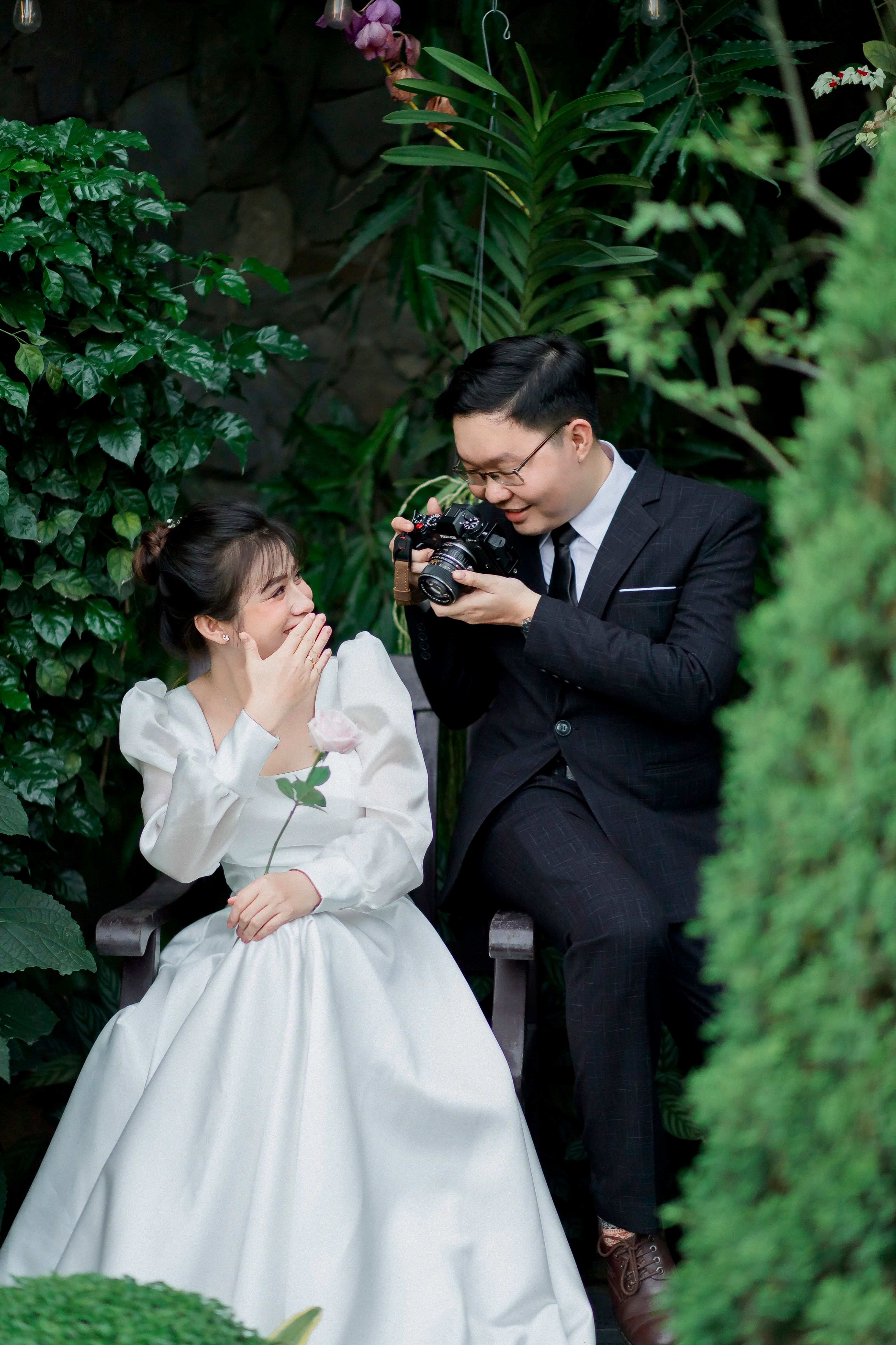 A groom playfully photographs his laughing bride in a lush garden setting.
