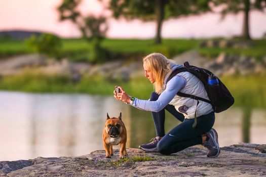 A woman photographing her French bulldog near a pond at twilight creates a serene outdoor moment.
