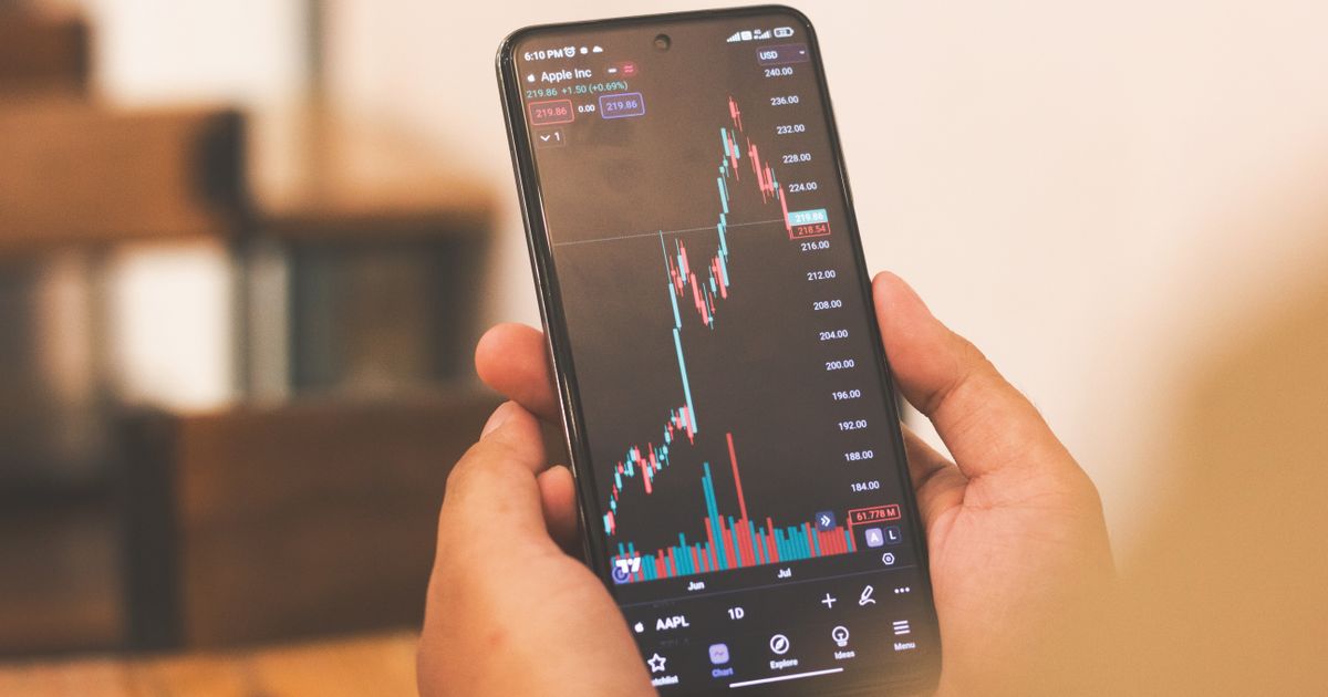Phone showing a stock candlestick chart on a trading app, held above a wooden desk