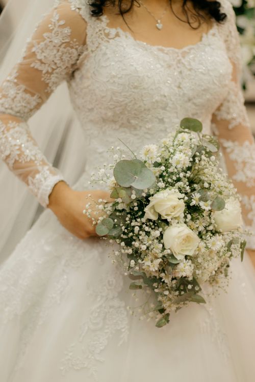 Free Close-up of a bride in lace gown holding a bouquet of white roses and greenery indoors. Stock Photo