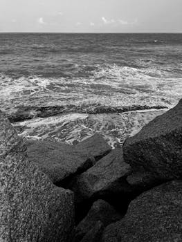 A serene black and white seascape featuring waves crashing on rocky shores during sunset.