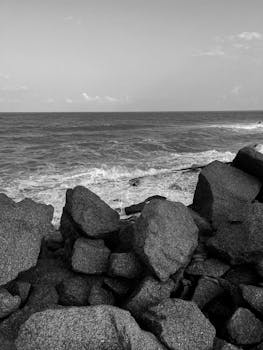 Black and white photo capturing rocky beach seashore with waves crashing at sunset.