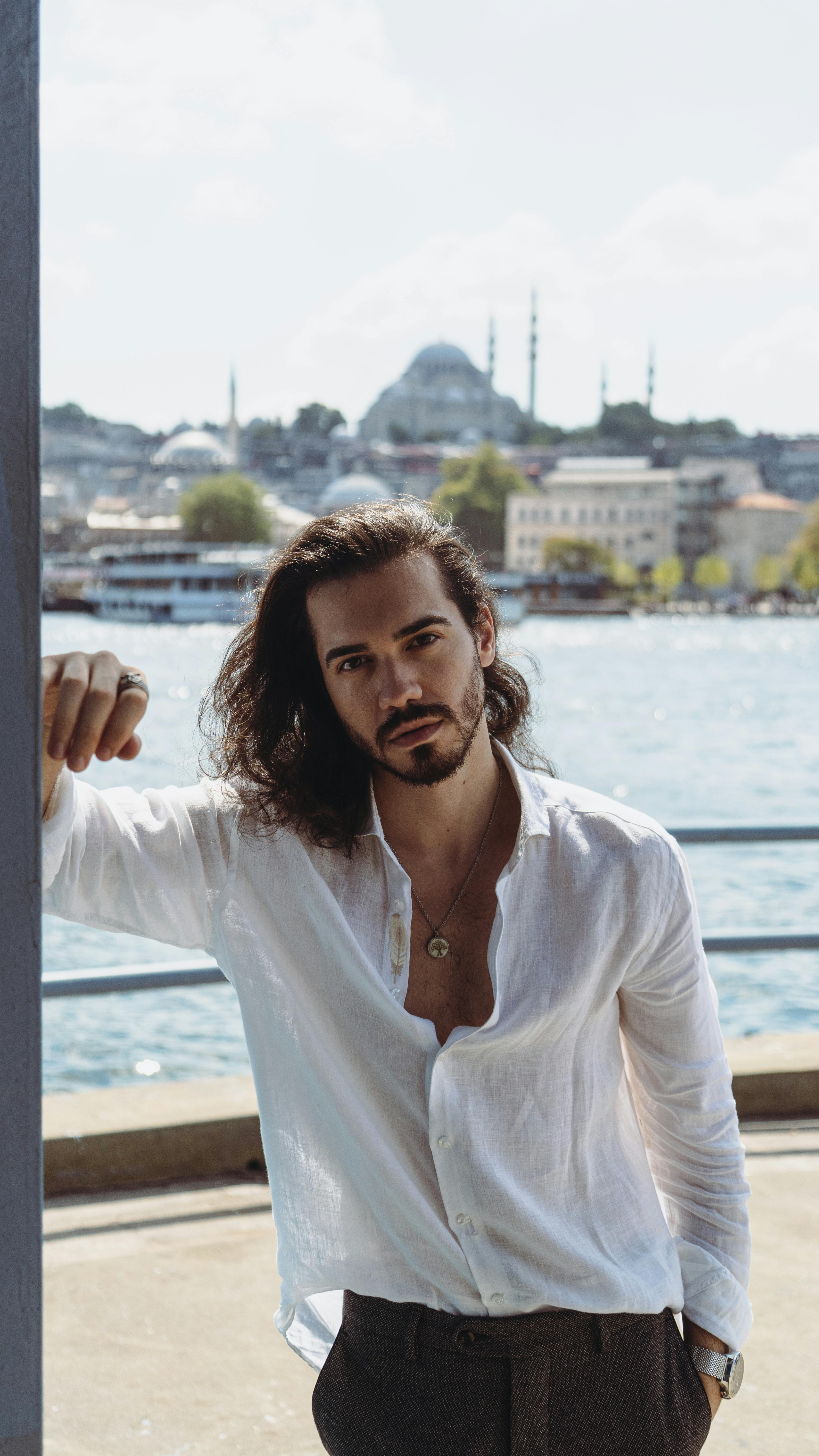 A young man in casual attire poses by the sea with a view of Istanbul's iconic skyline.