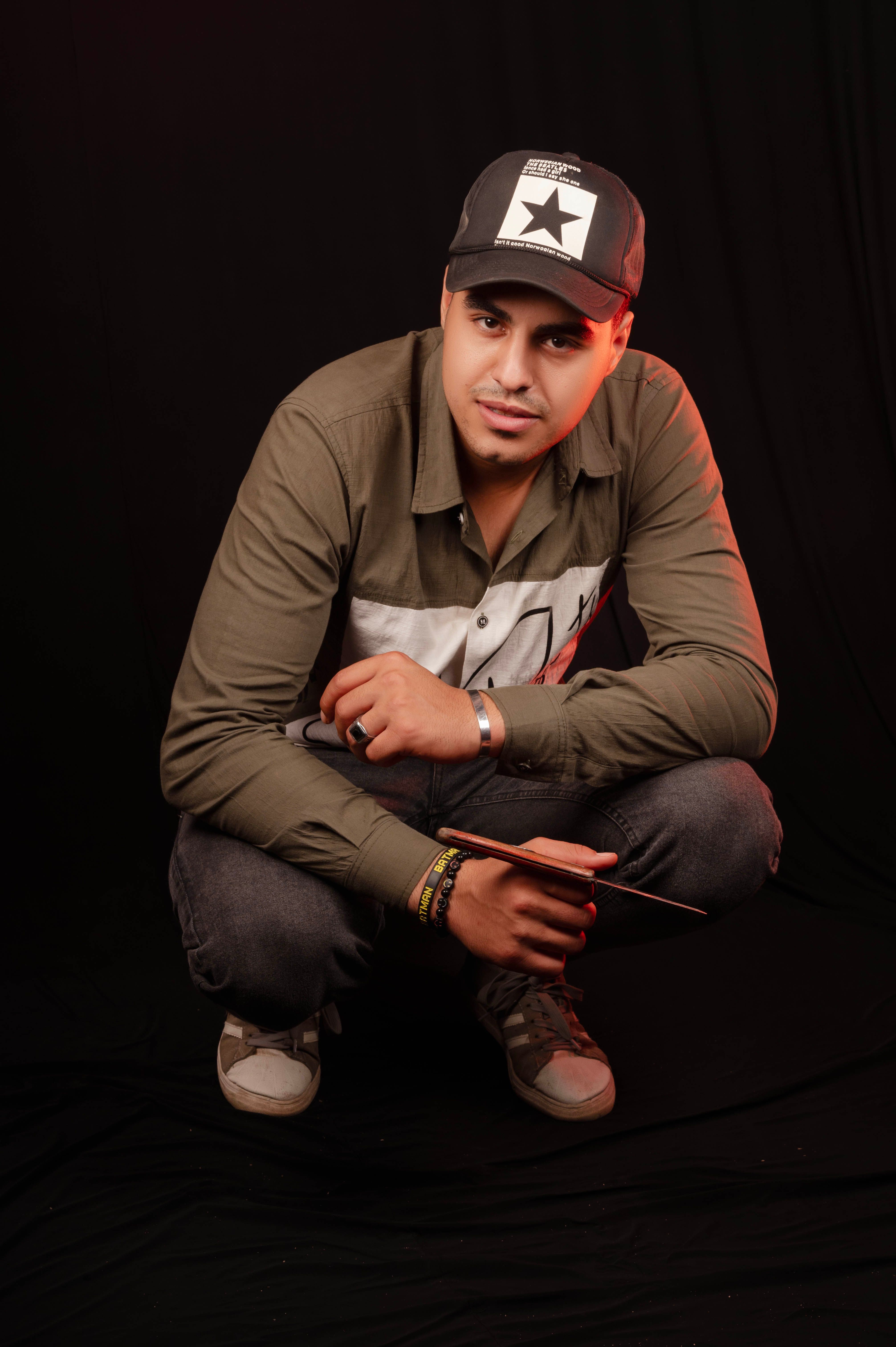 A man poses playfully in a studio, wearing a casual outfit and cap, with a dark backdrop.