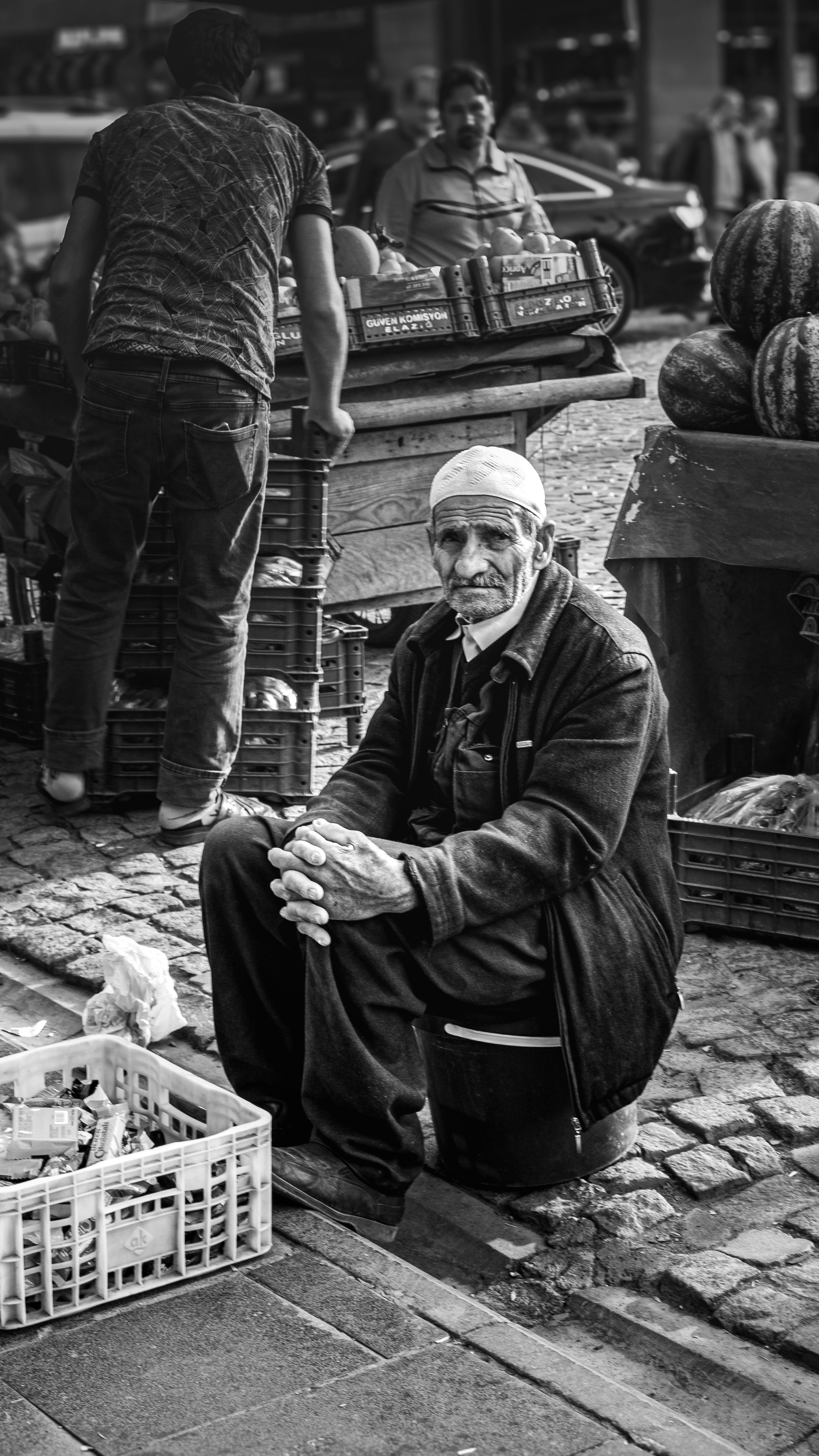 Black and white street portrait of an elderly merchant in a busy market surrounded by produce.