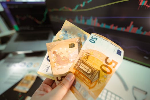 Hand holding Euro banknotes in front of a trading screen showing financial graphs.