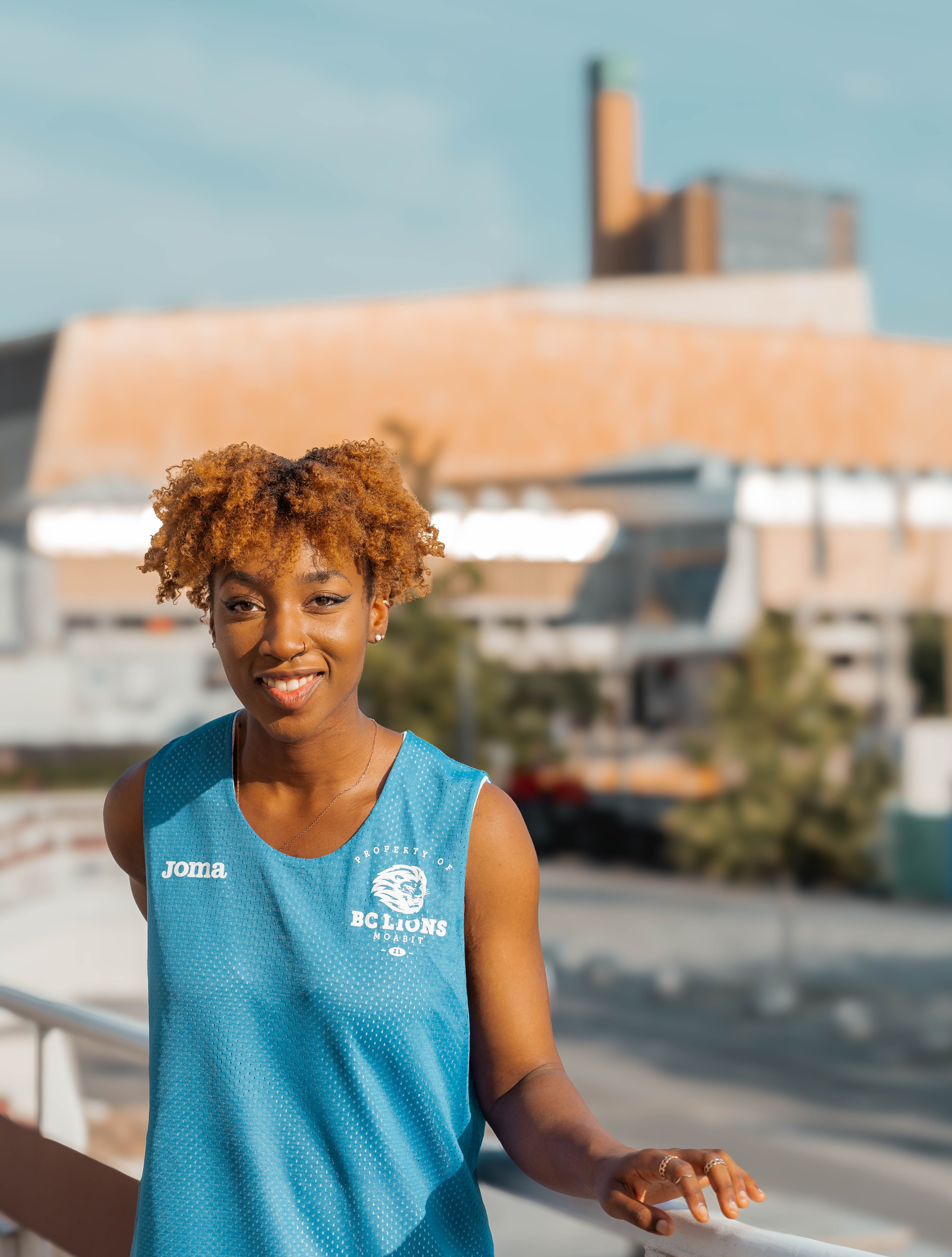 Portrait of a smiling athlete in a blue jersey standing outdoors on a sunny urban balcony.