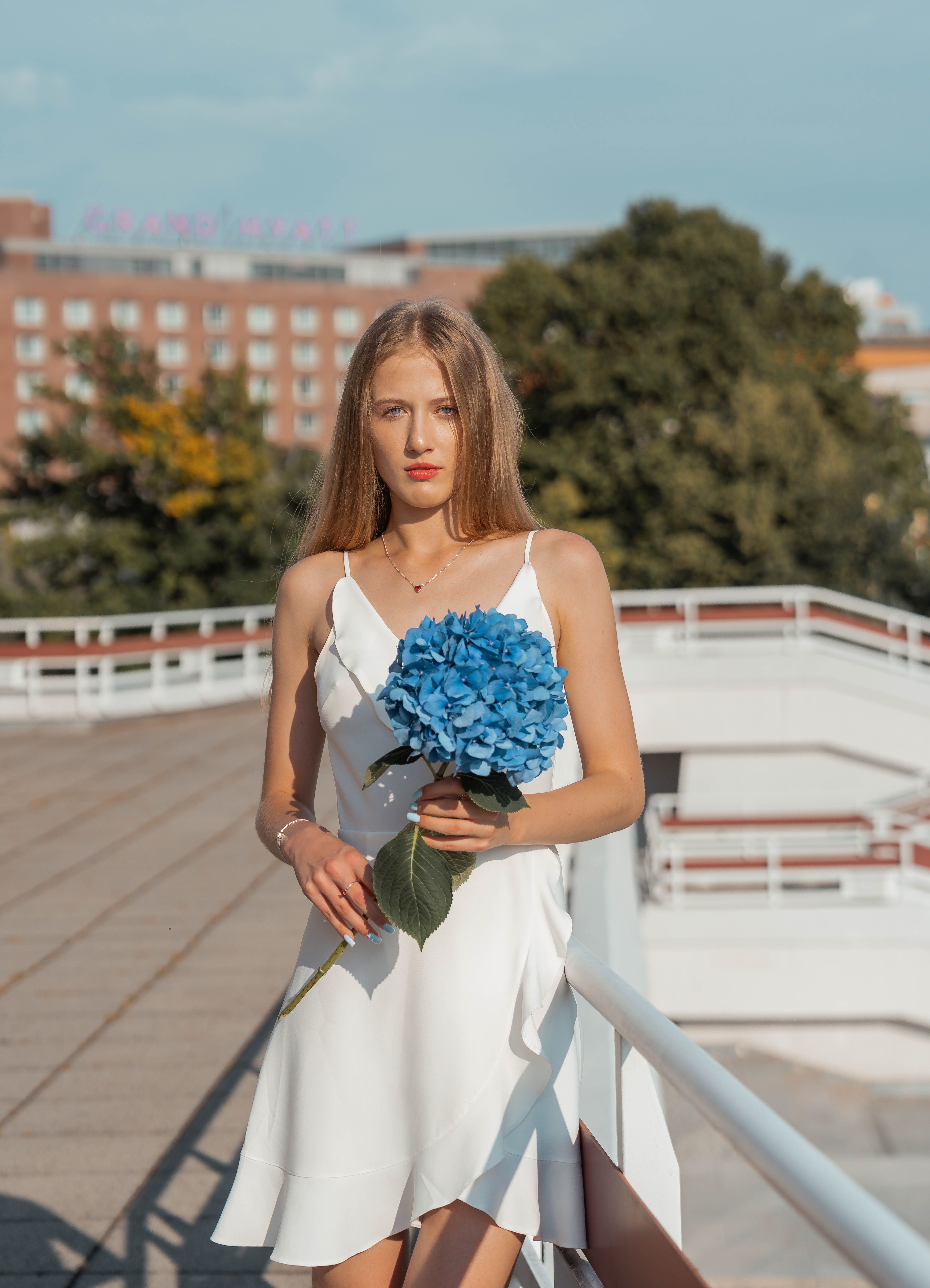 Portrait of a stylish woman in a white dress holding a blue hydrangea flower, captured outdoors in a sunny setting.