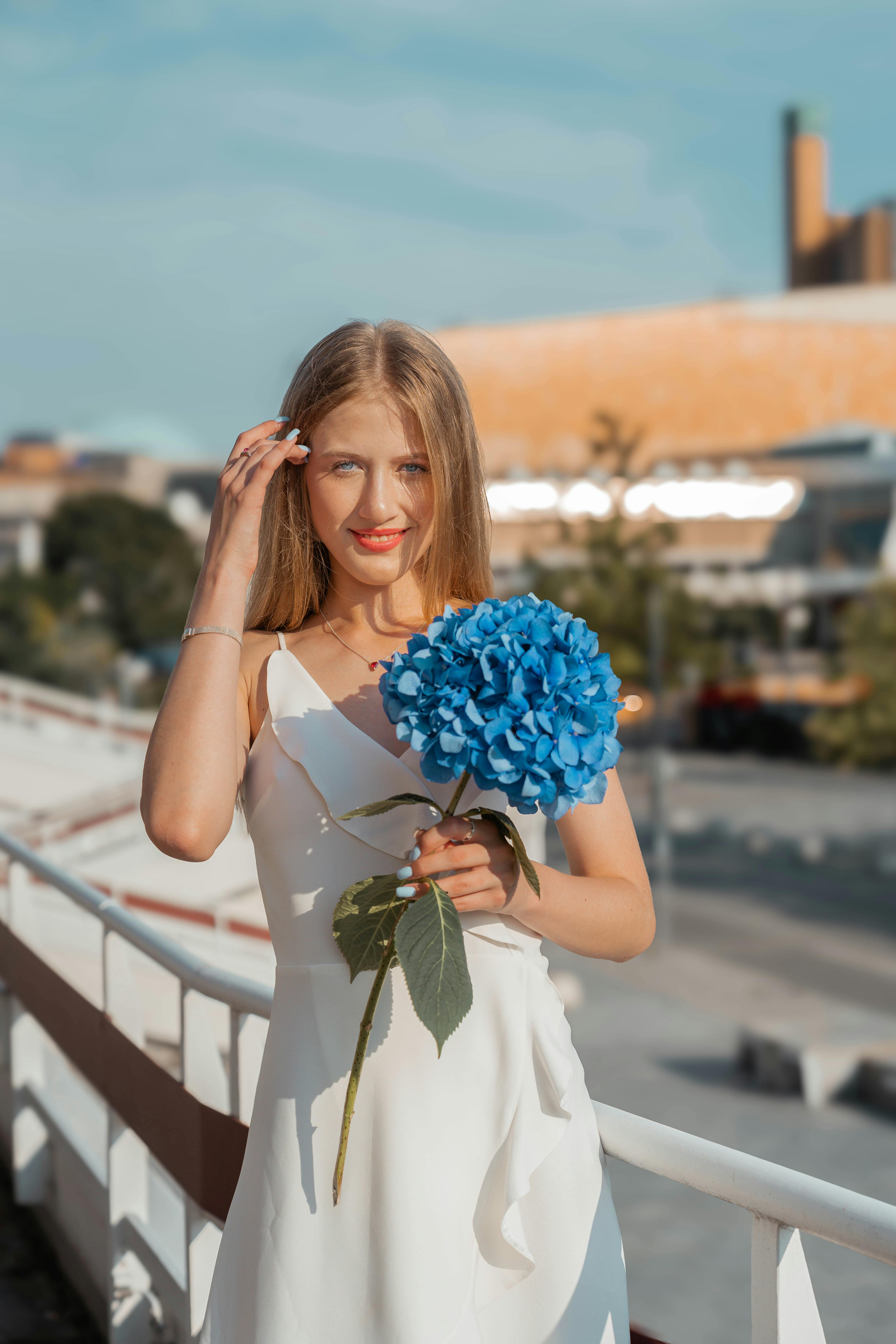 A woman in a white dress holds a vibrant blue hydrangea with a bright summer backdrop.