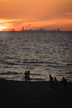 Silhouetted people walking on a beach with a city skyline at sunset.