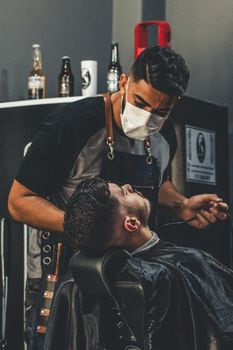 A barber wearing a face mask gives a haircut to a client in an indoor barbershop setting.