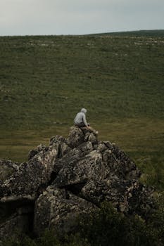 A solitary individual sits atop a large rock formation surrounded by expansive green fields under a cloudy sky.