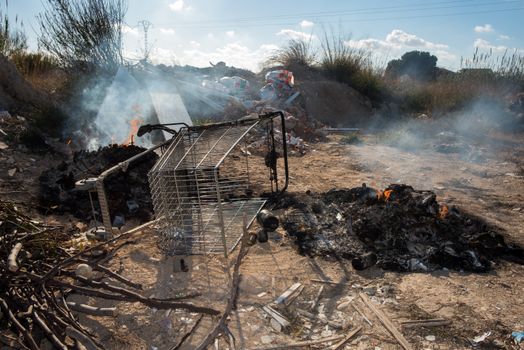 A shopping cart amidst waste and smoldering debris in an outdoor landfill, conveying environmental neglect.