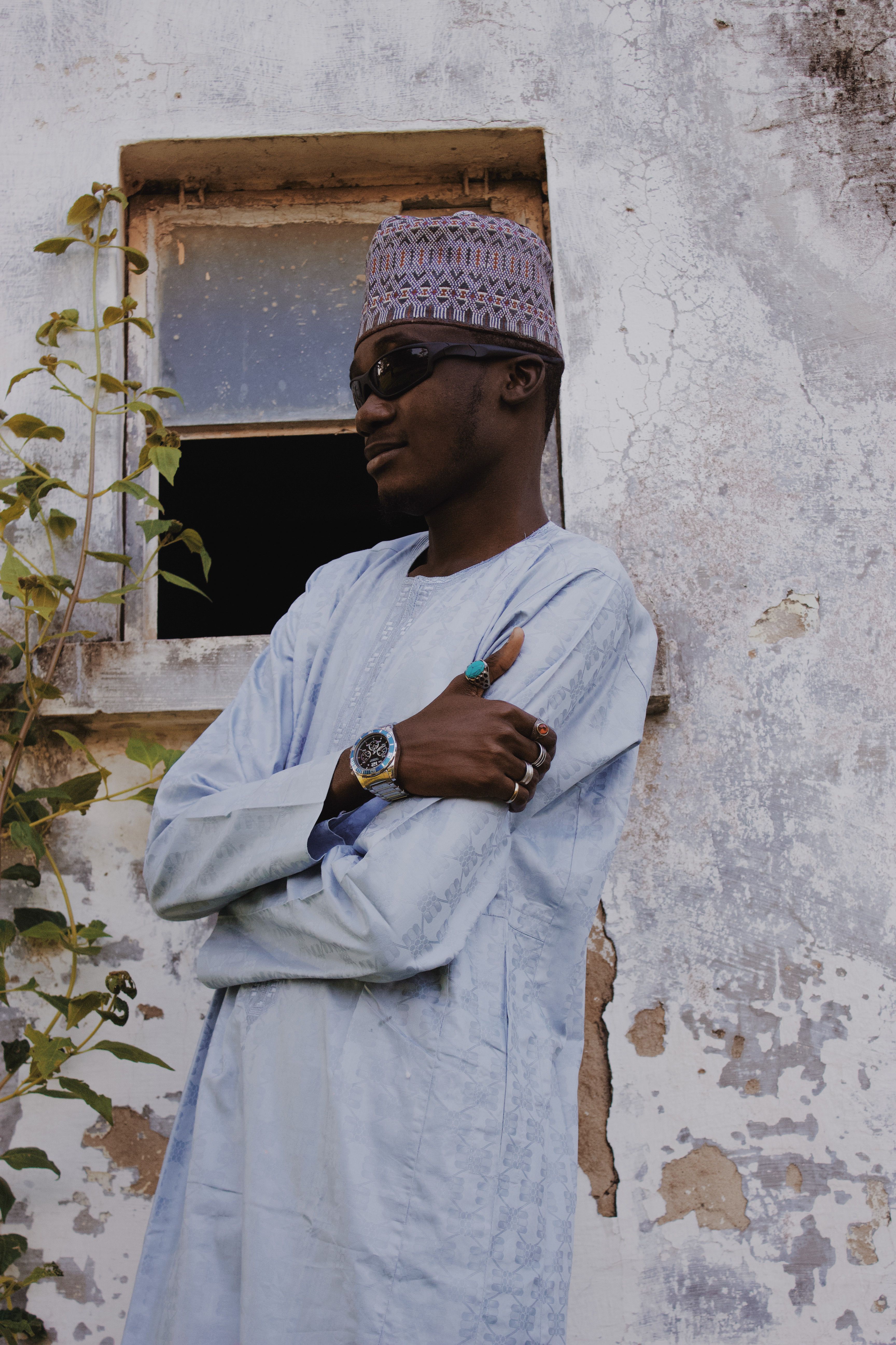 Man wearing traditional attire and sunglasses stands against an old wall.
