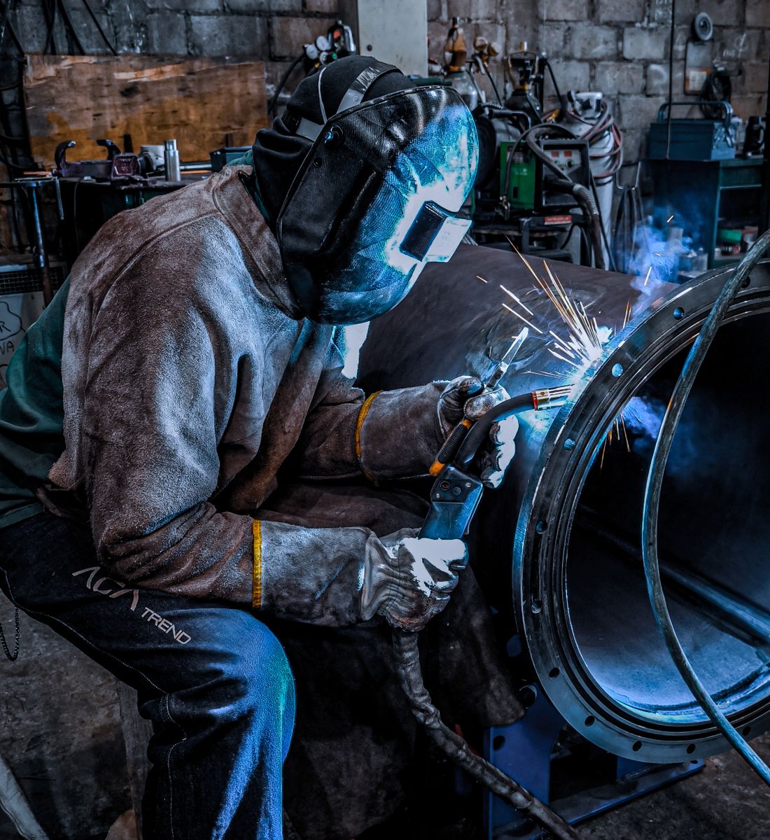 Welder in a protective helmet and leather gloves welding a steel component inside a workshop.
