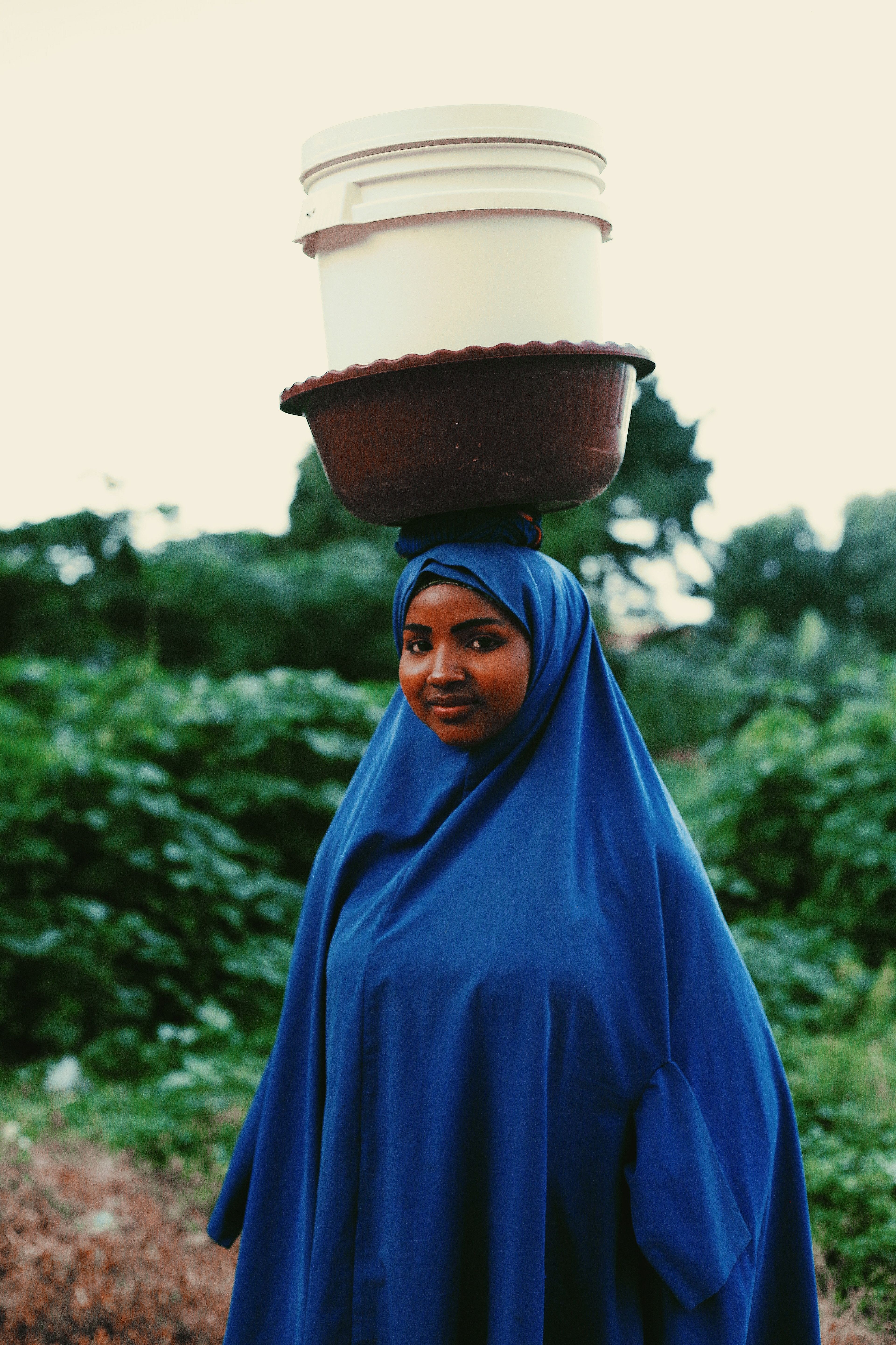 A woman in blue hijab carrying a bucket on her head · Free Stock Photo