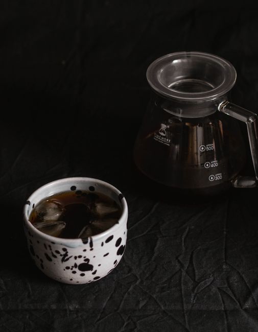 A minimalist composition featuring iced coffee in a ceramic cup and a glass carafe on a dark textured surface.
