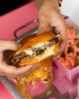 Close-up of hands holding a juicy cheeseburger with a pink background.
