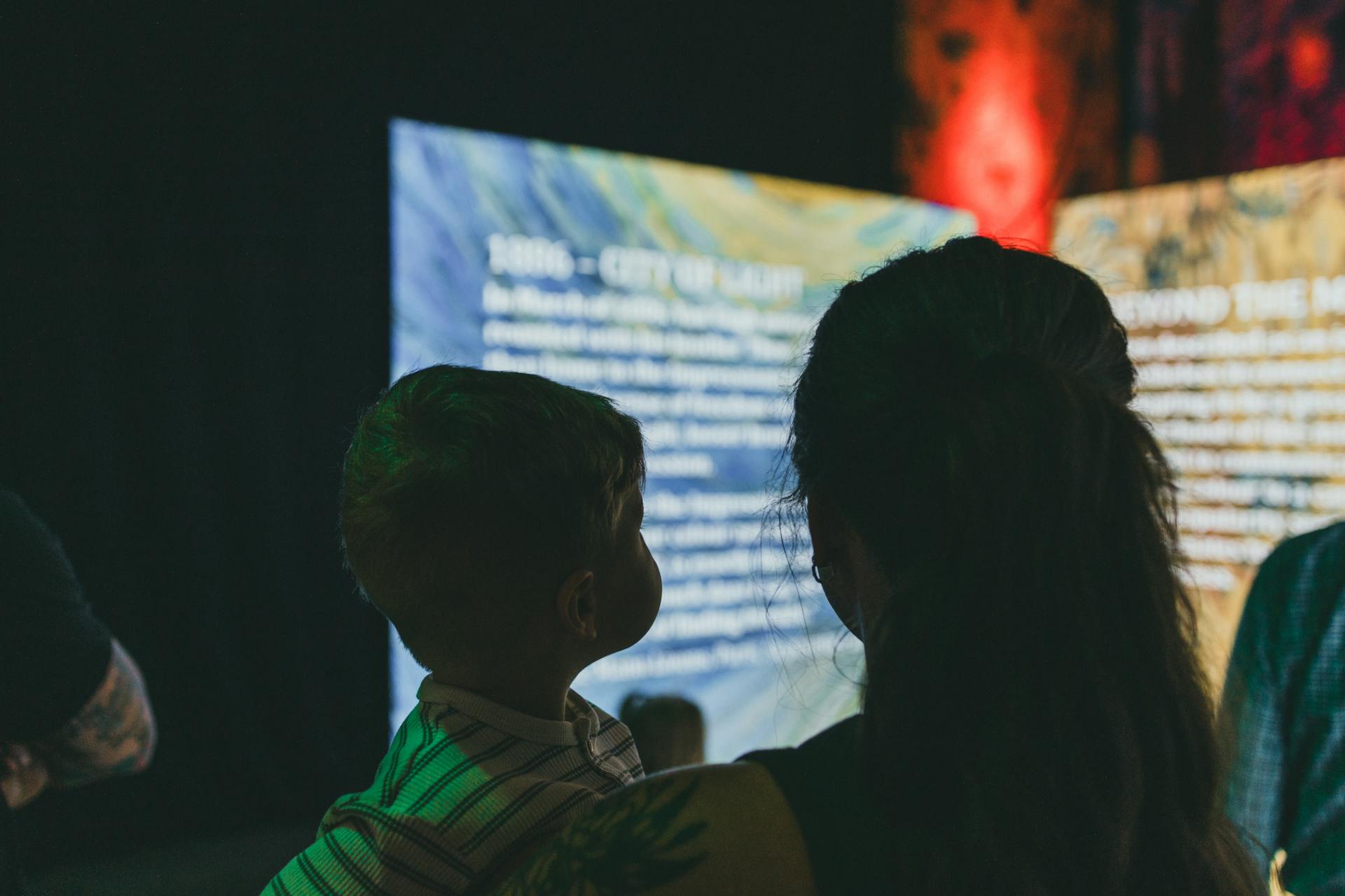 Silhouettes of a woman and child observing digital art and text on illuminated screens indoors.