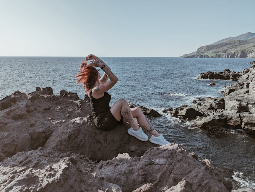 A woman with tattoos enjoys a peaceful moment on the rocky coastal shore, basking in the summer sun.