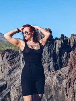Young woman with tattoos posing confidently by rugged rocks under clear sky.